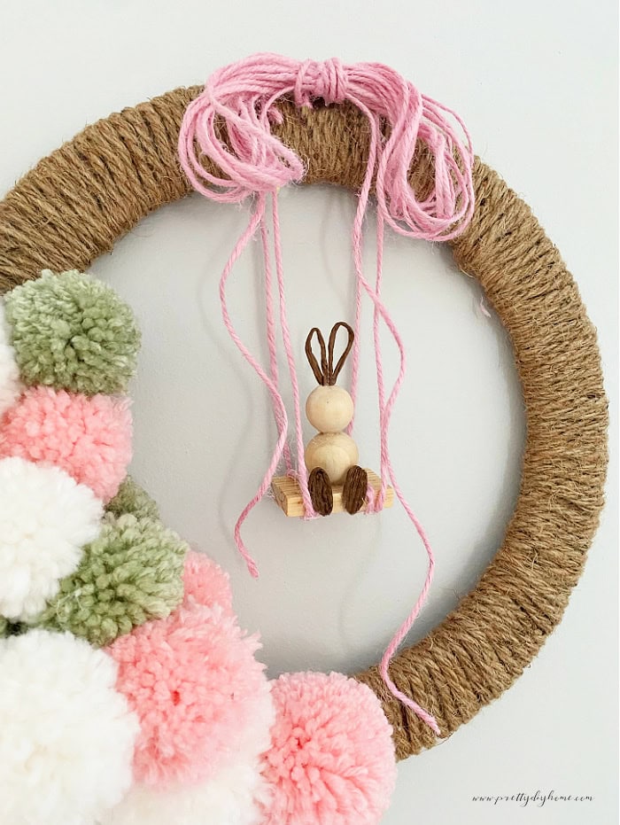 A diy Easter Wreath craft made with wrapped twine, a dozen pastel colored pom poms and a DIY wood bead bunny hanging from a swing as a centerpiece.