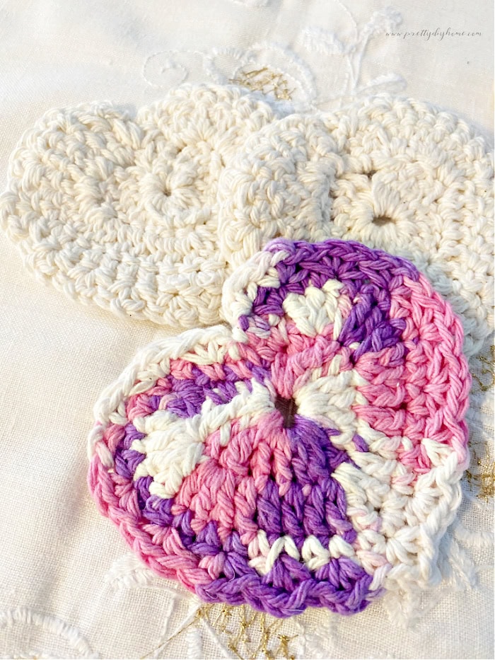 Three crocheted scrubbies made with cotton yarn. Each crochet scrubby is heart shaped, two are white and one is purple pink and white.