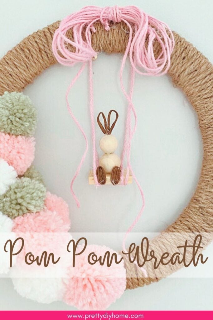 A diy Easter Wreath craft made with wrapped twine, a dozen pastel colored pom poms and a DIY wood bead bunny hanging from a swing as a centerpiece.  The overlaying text says pom pom wreath.