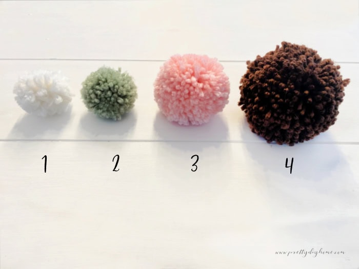 Four different sized DIY pom poms made with a Clover pom pom maker.  They are each in a different color, white, green, pink and brown.