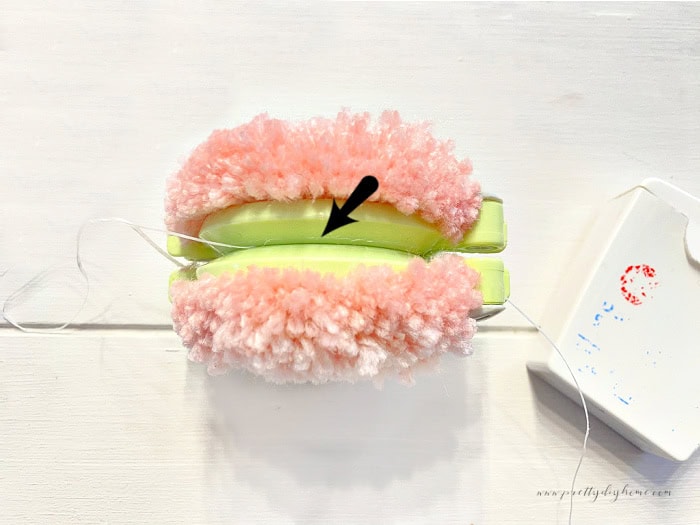 Tying the center of a pom pom with dental floss.   There is an arrow showing the placement of the tie into the groove on the maker.