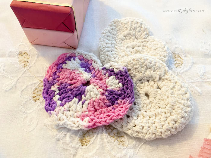 Three homemade crocheted face scrubbies in the shaped of Valentines.  The three heart shaped face scrubbies are laying on top of a white vintage cloth with a stack of strawberry scented soaps beside them.  Its a great DIY Valentine's gift idea.