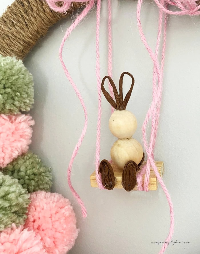 A miniature Easter bunny made with wood beads and wire ears. The little bunny is sitting on a swing made out of jenga wood blocks from Dollar tree and paper covered wired. The side of the image shows pom poms that are part of the wreath.