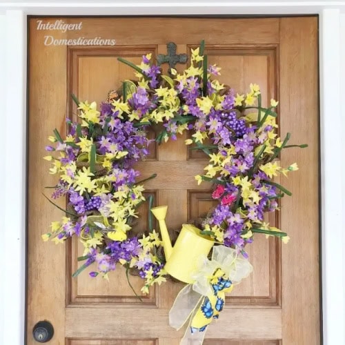 A large and lush Spring wreath made with bright yellow and purple flowers.  The diy wreath is embellished with a yellow watering can and pretty yellow ribbon.