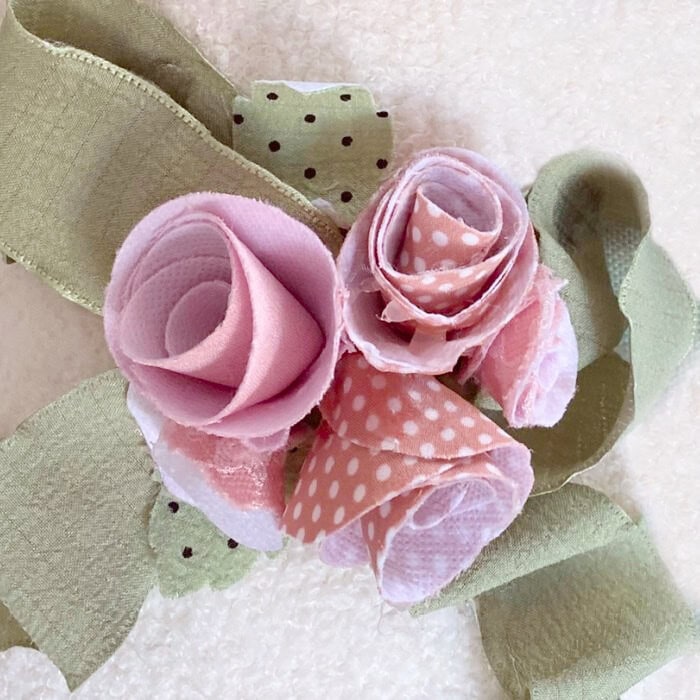 Three rolled fabric flowers made with soft pink and pink polka dot fabric, the three diy flowers are compliments with soft sage green ribbon and diy fabric leaves.