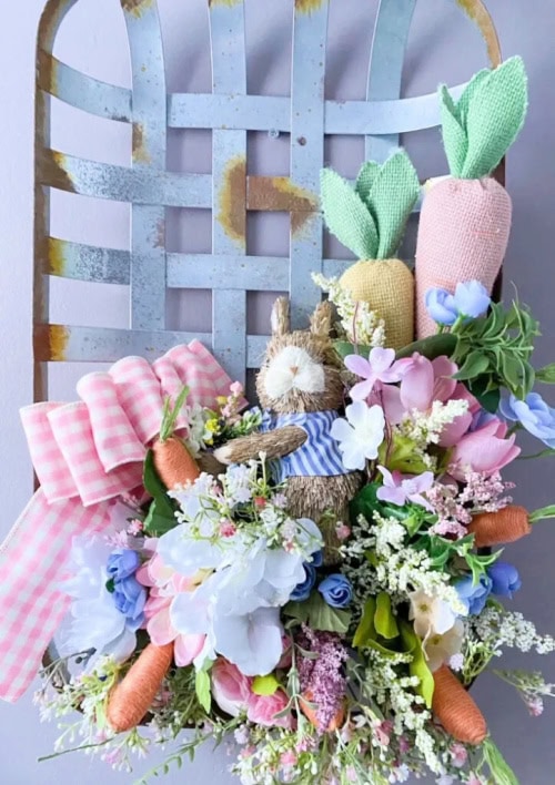 A very colorful DIY Spring wreath made using a tobacco basket, a small stuffed wicker Easter bunny, stuffed carrots and assorted brightly colored blossoms.