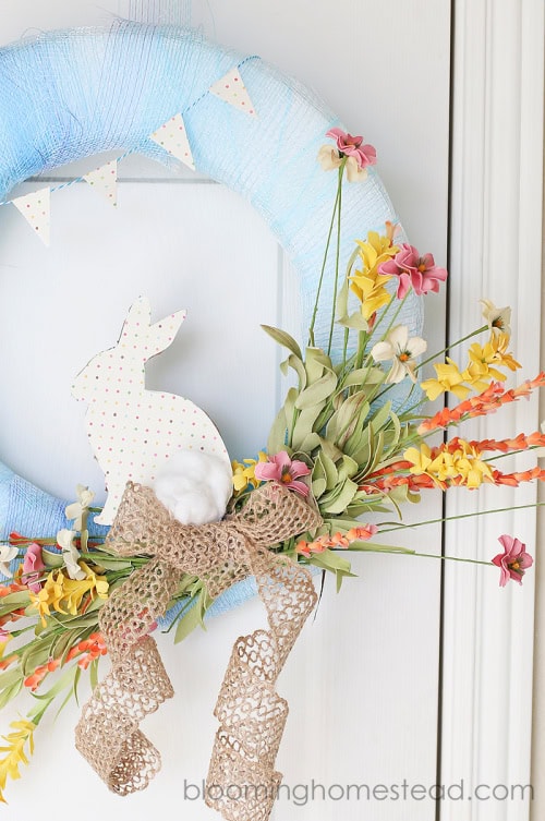 A gorgeous blue wreath form is the highlight of this beautiful DIY Easter wreath.  There is a polka dot bunny with matching banner hanging across the wreath and its embellished with bright pink and yellow florals and finished with a burlap bow.