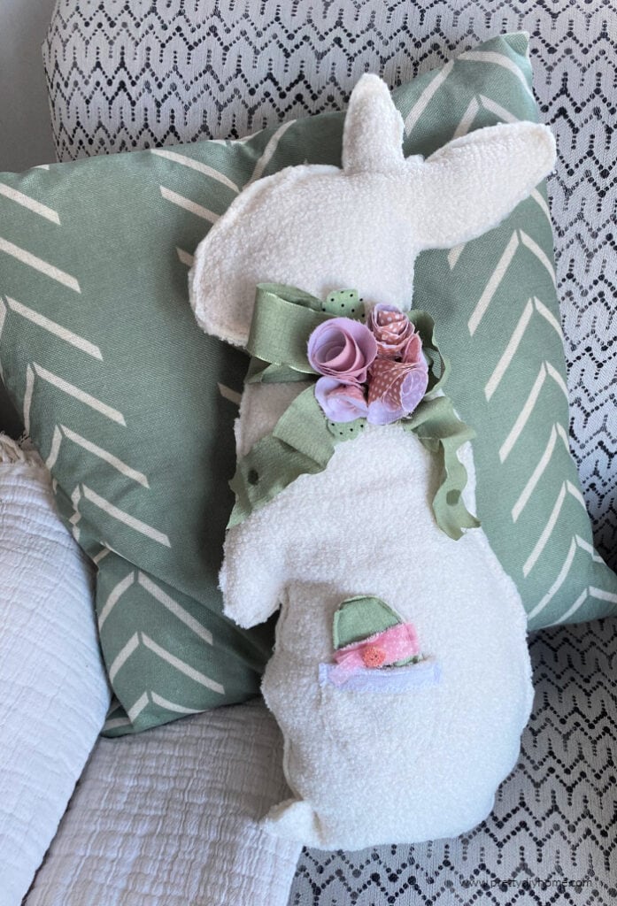 A white fluffy bunny cushion decorated with pink fabric flowers and green ribbon. The bunny shaped cushion is sitting beside a green cushion,