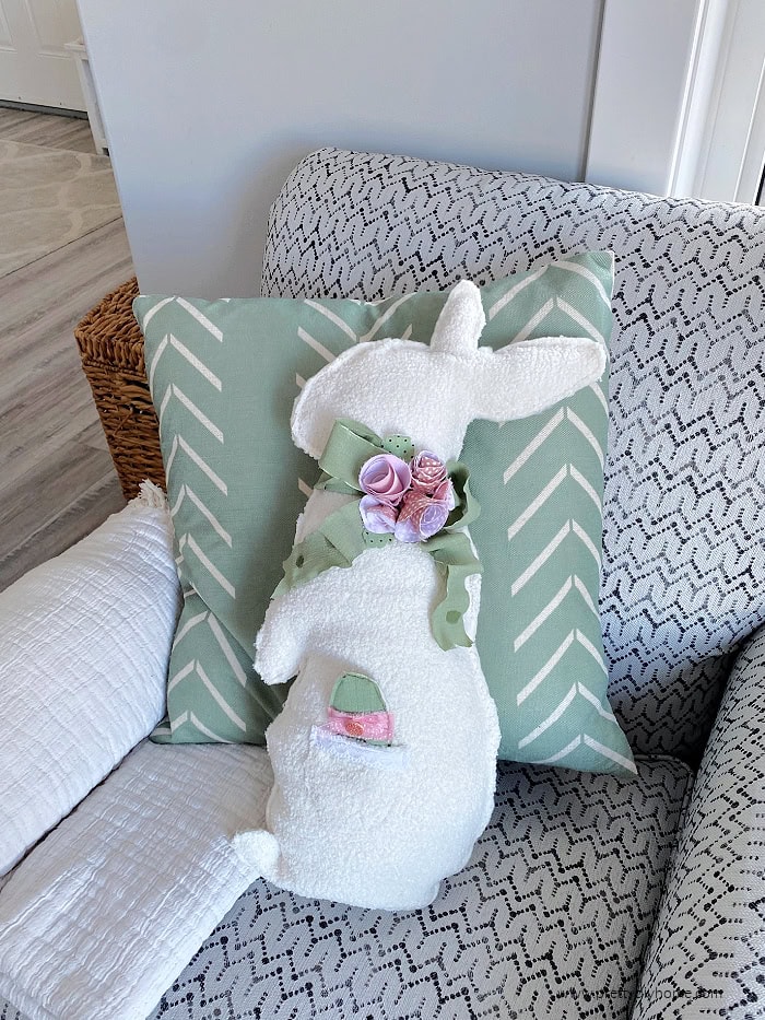 A livingroom chair decorated for Easter with an adorable bunny shaped cushion, The bunny cushion is sew from white woolish Dollar Tree fabric and decorated with a pocket holding a stuffed Easter egg. The neck is decorated with soft green ribbon bow, and rolled fabric flowers in different shades of pink.