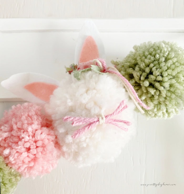 A fluffy white Easter bunny pom pom craft in soft white yarn, with pink and white ears made with felt. Their is pink whiskers made with twine, and its decorated with beautiful fabric flowers near the ears.