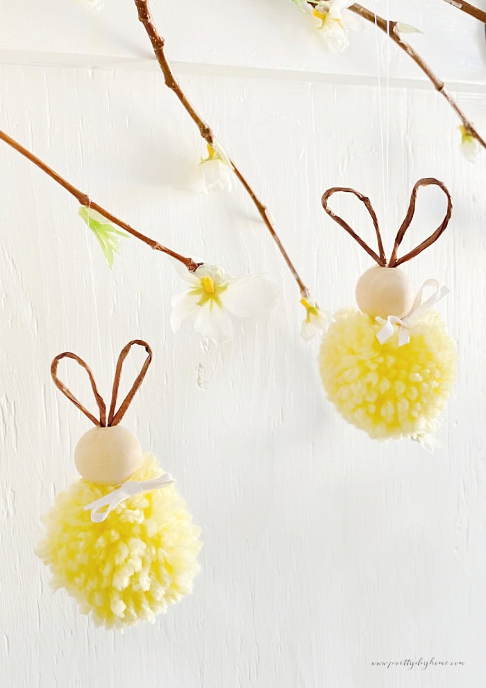 Two yellow pom pom bunnies hanging from a Easter branch with yellow and white flowers.