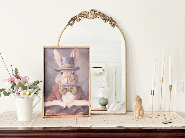 Easter decor arrangement on a sideboard featuring a large DIY canvas frame with an easter bunny art canvas. There is also a bouquet of flowers, some tapered candles, and a large gold mirror as the background.