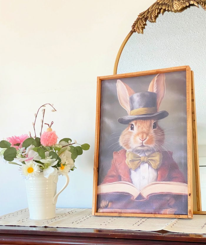 A livingroom side table decorated with a mirror, rabbit painting and a floral arrangement thats been embellished with a pom pom bunny ornament