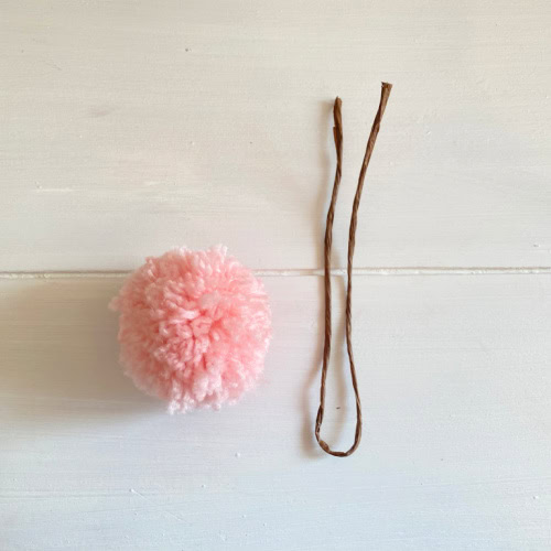 A small pink pom pom and a piece of folded paper covered wire for a tutorial on how to make a yarn Pom Pom bunny for Easter.