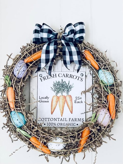 A dollar tree fresh carrots sign is the backdrop to a colorful  DIY Spring wreath.  The wreath is decorated with a double layer of wire wreath forms stuffed with easter grass, speckled easter eggs and tiny orange carrots.