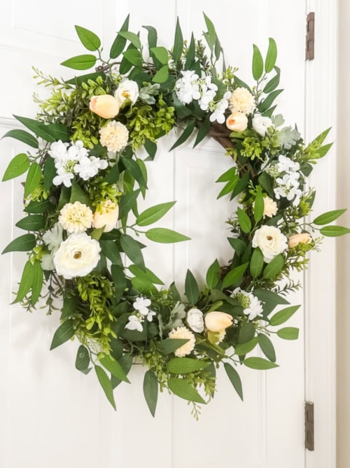 A large and luscious green floral wreath with lots of thick dark greenery, softer green sprigs, then topped with small yellow tulips, mums, and then small white florals.