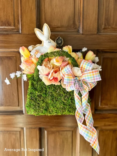 A DIY Spring wreath craft idea made using a moss purse and the foundation. Add some pretty peach and white florals, and a large white bunny finished off with a multicoloured checkered ribbon.