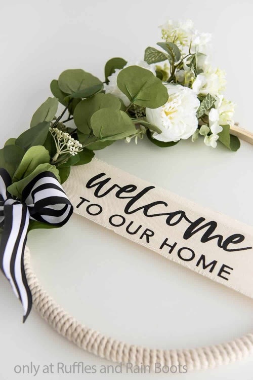 A neutral white and green embroidery hoop wreath for Spring.  The wreath has welcome to our home written across the middle using a cricut.