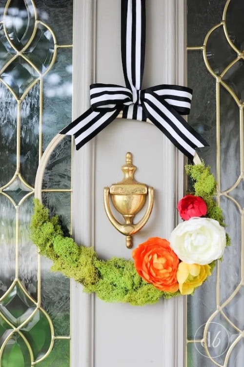 A cheerful and brightly colored DIY Spring wreath made using an embroidery hoop, bright green moss, and orange and white flowers.  The wreath is hanging on a white front door, and is hung with a black and white dramatic striped ribbon.