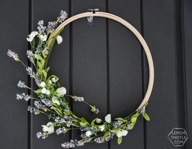 An simple diy Spring wreath made with an embroidery hoops and half of the hoops is wrapped with white florals with greenery and small lavender flowers. Its dramatic hanging on an dark black door.