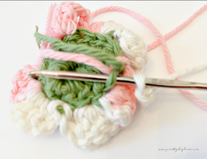 The backside of a pink white and green crocheted flower.  Their is a crochet hook inside a back loops of the crochet project illustrating stitch placement.