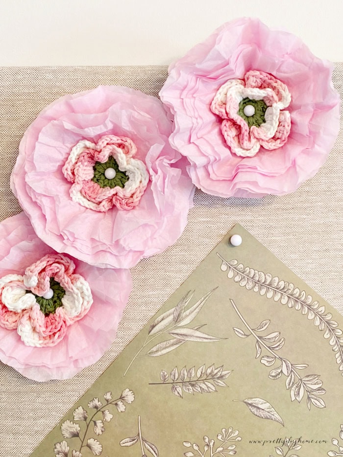 A closeup of three pretty pink handmade crocheted flowers attached to a bulletin board for craft room decor.