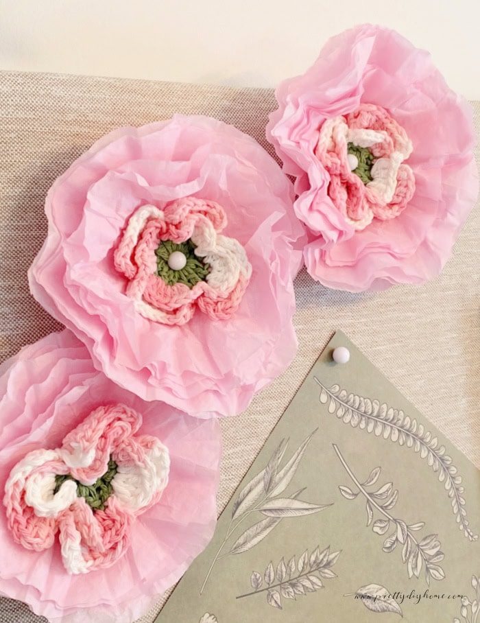 A closeup of three beautiful crocheted flowers, surrounded by coffee filter petals to make the flowers larger. The center of the flowers are green the crocheted part is white and pink and the outside coffee filter petals are a soft pink.