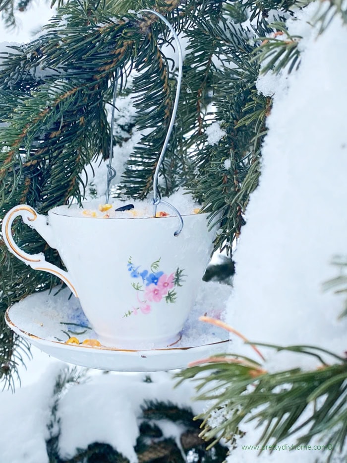 A suet teacup bird feeder hanging in a evergreen tree thats still covered in snow. The diy teacup bird feeder is white with flowers and a sweet gold handle.