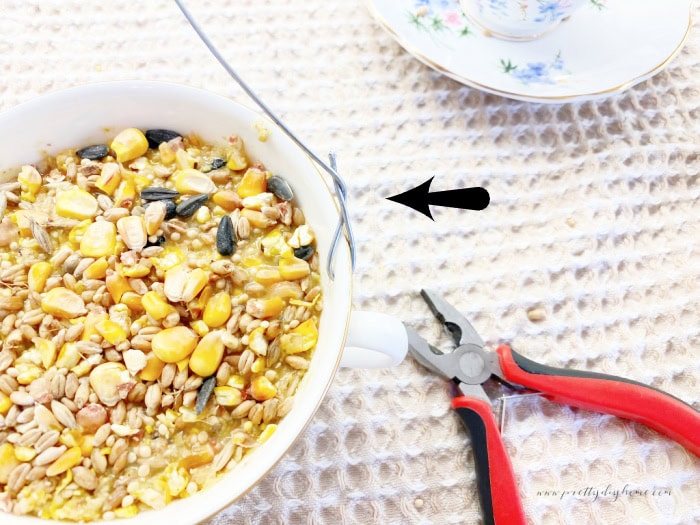 A tea ccup bird feeder filled with suet and bird seed.  There is a set of plyers beside the teacup and an arrow pointing to how the wire is wrapped for hanging from the tree.