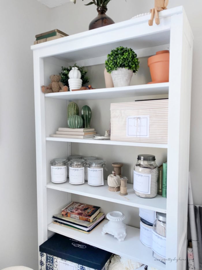 A craft room storage shelf filled with pretty ornaments, craft room storage jars with labels, a stack of wool, and a matching storage box.