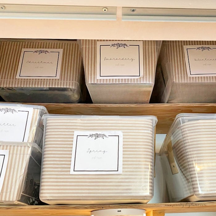The upper shelves of a craft closet filled with pretty storage bins of  craft supplies.  Each bin is lined in matching paper with pretty matching labels identifying whats inside.
