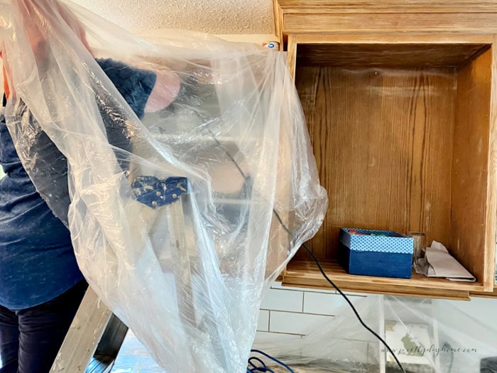 Kitchen cupboards being sanded with a woman on a ladder underneath plastic to reduce dust.