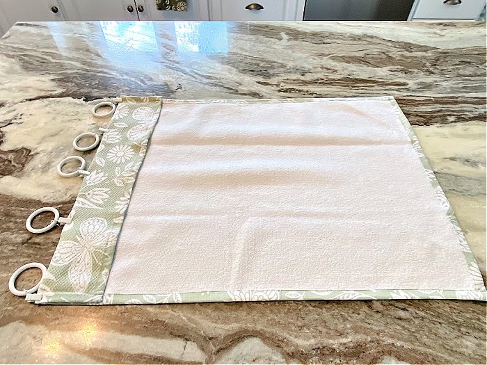 A DIY kitchen curtain laing flat on a counter. You can see where it is folded, and clipped at the top to make cafe style kitchen curtains.