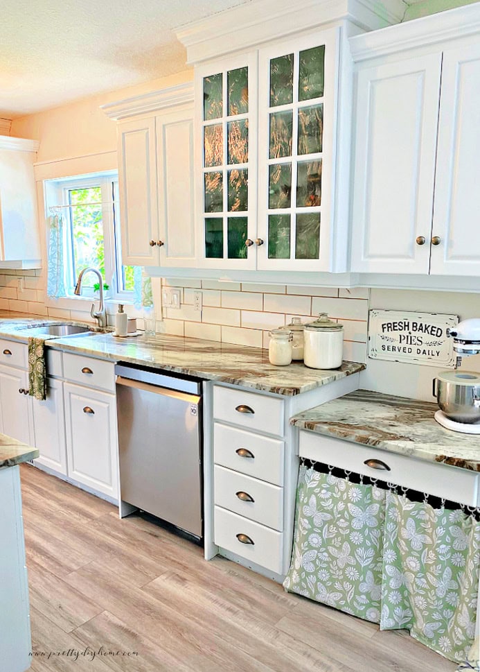 A kitchen with white cabinets and a green kirchen window curtains. The kitchen half curtains are hanging over the sink and as a skirt on the lower cabinets.