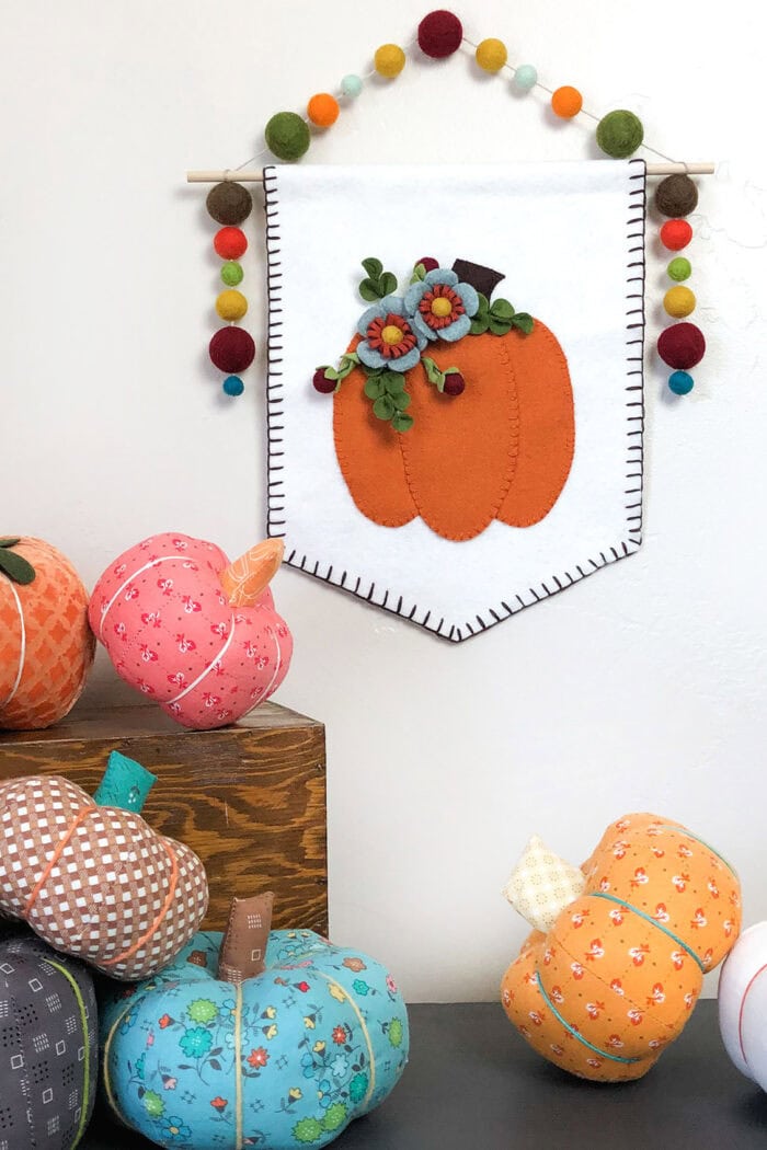 A beautiful large banner hanging from a dowel in white fabric.  The fabric is decorated with a beautiful felt hand stitched pumpkin that is embellished with felt flowers.