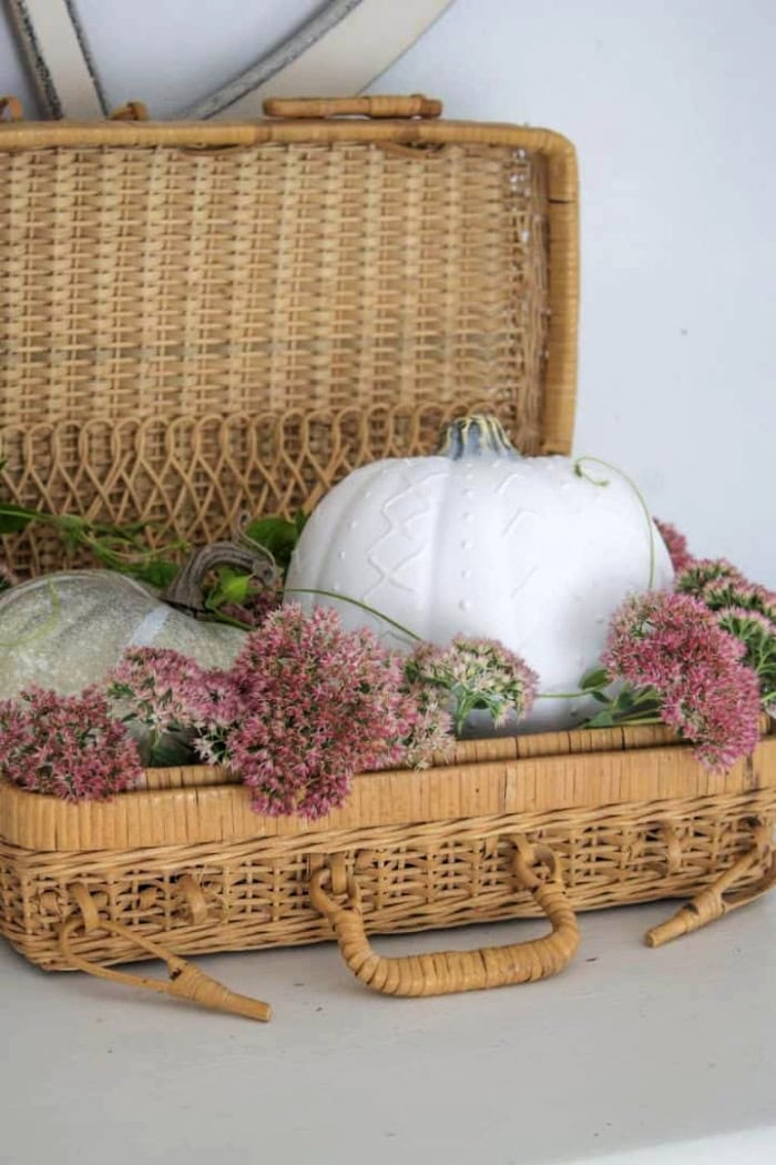 A pumpkin painted white and decorated to look like its a arglan sweater using puff paint. The pumpkin is sitting in a vintage basket with a lid and surrounded by pretty pink fall flowers.