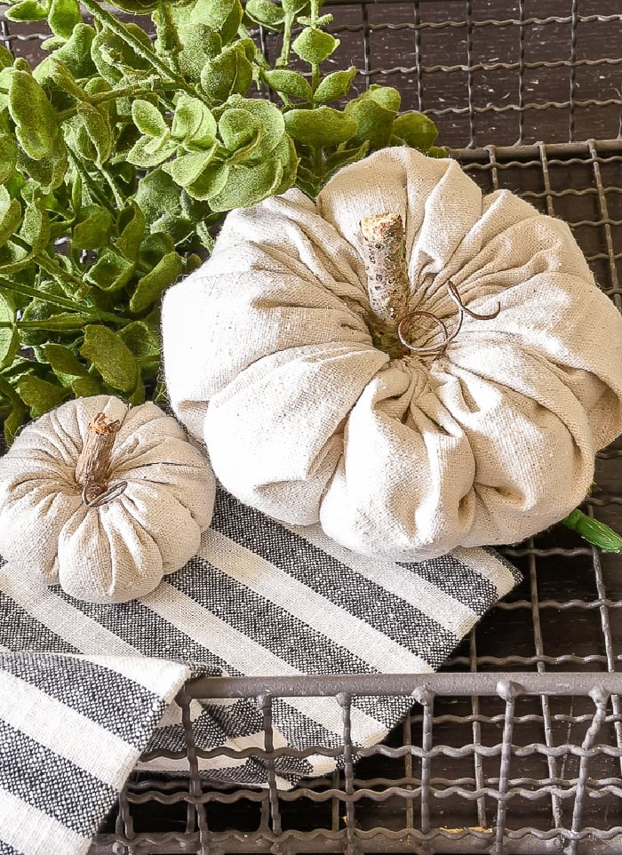 Neutral  colored fabric pumpkins in a soft cream colored fabric.  The two diy fabric pumpkins are sitting in a basket with a striped cloth and greenery,