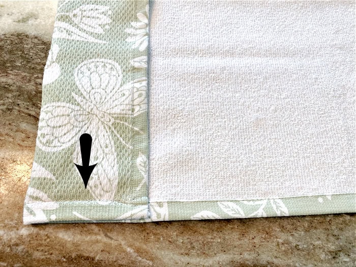 A kitchen towel being used to make easy sew curtains. Top of the kitchen towel is folded over and there is an arrow pointing to the short seam on the side. The kitchen curtains can also be made no sew using iron on stitch or glue.