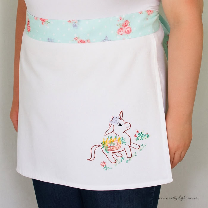 A woman wearing a handmade half apron using a tea towel. It has cute donkey embroidery on the corner .