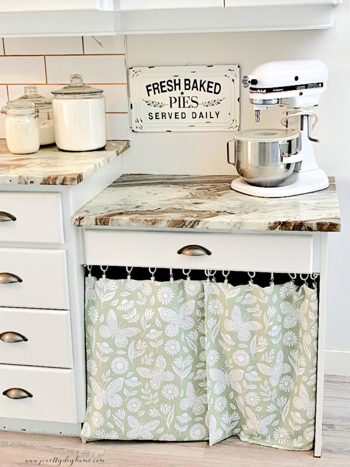 A set of easy kitchen curtains hanging on lower cabinets in soft green. The curtains add a soft cottage style vibe to the white kitchen cupboards.