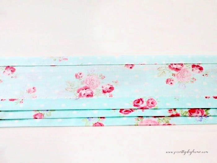 Three strips of fabric cut and ironed for making an apron. The three strips of fabric are in a very pretty cottage style fabric with baby blue cotton fabric and pink flowers.