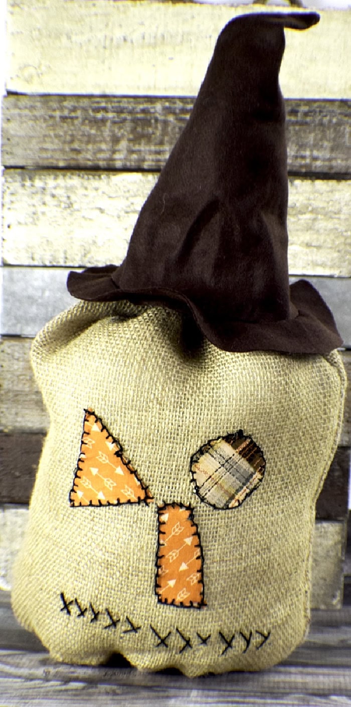 A fun scarecrow pumpkin the pumpkin is made with burap decorated with sewn on fabric patches for a face and feature a dark brown hat.