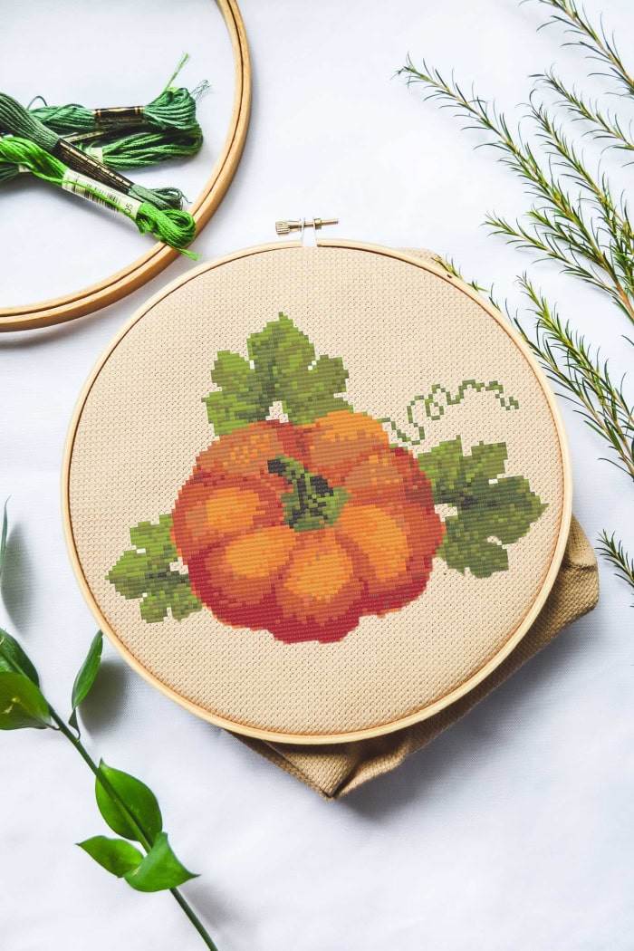A cross stitched pumpkin craft in orange and green done on cream colored alda cloth.