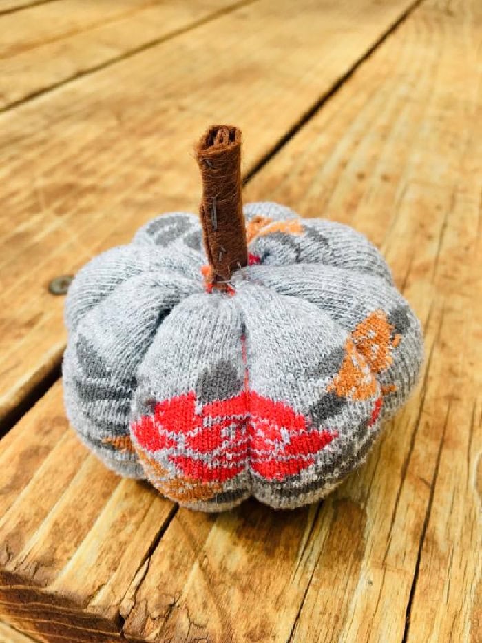 A sock pumpkin sitting on a wooden table. The pumpkin sock has orange and red pattern woven into it.