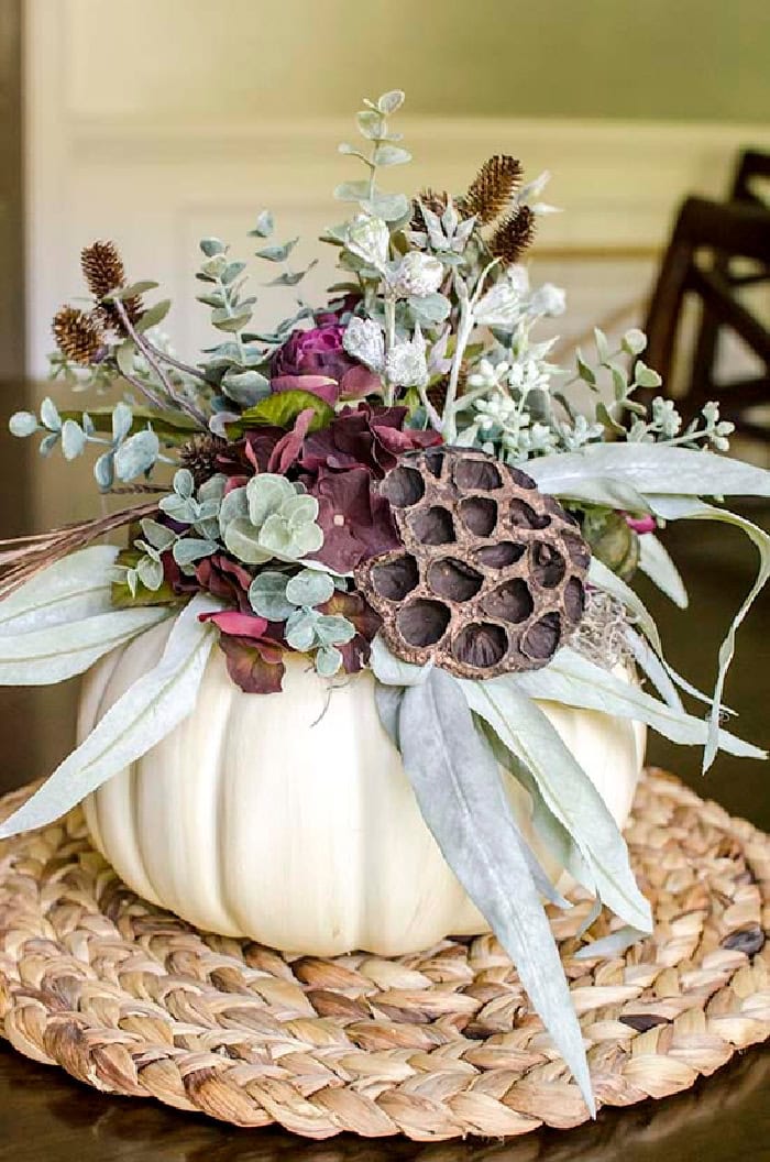 A large pumpkin centerpiece with a white pumpkin decorated on top with assorted purple flowers, brown dried picks, and soft green leaves.