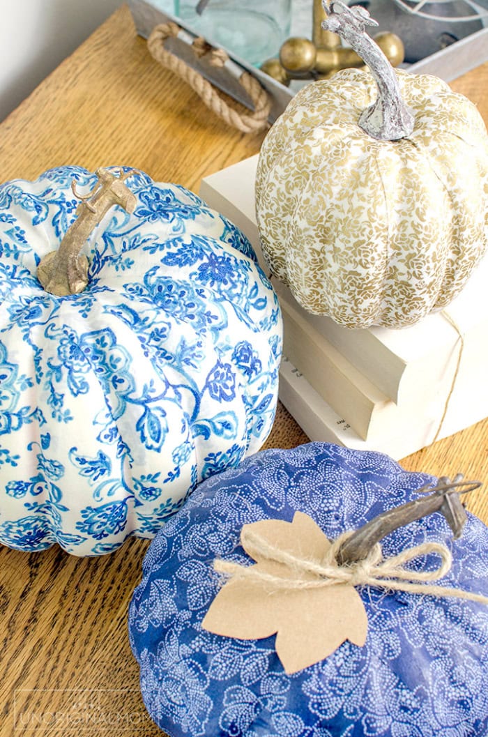Three fabric covered DIY pumpkins. One of the handmade pumpkins is dark blue with white, a second one is white with dark blue patternm and the third one is white a gold. These handmade pumpkin crafts are cheerful and bright sitting on an wood tabletop.