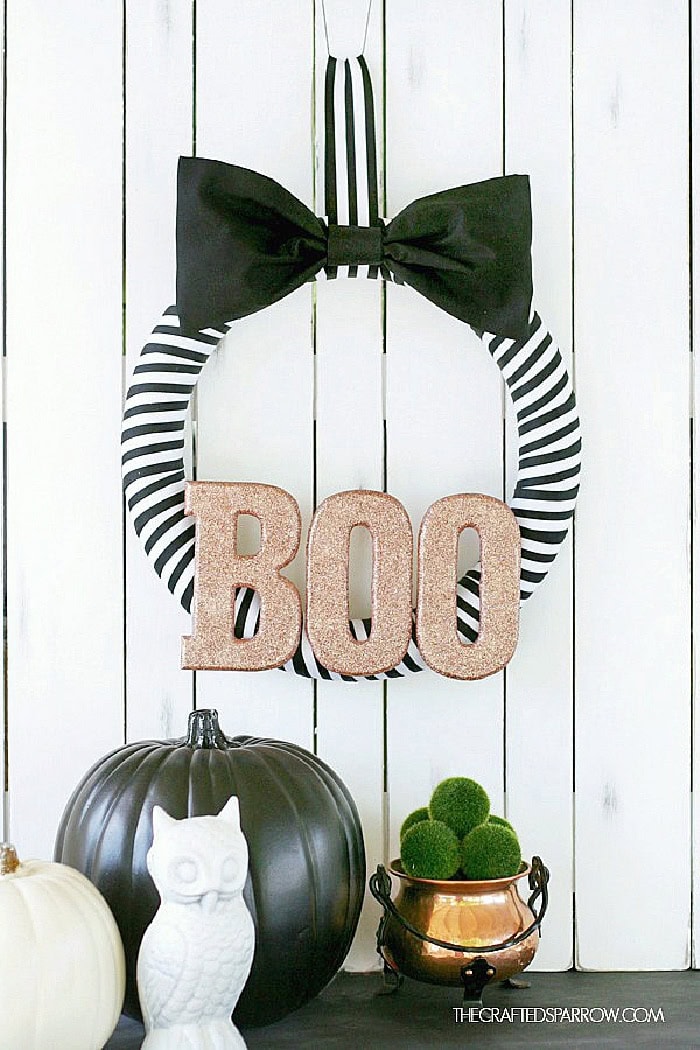 A minimalistic black and white DIY Halloween wreath with black and white stripped ribbon around a wreath form, a black bow on top and gol sparkly BOO at the bottom.