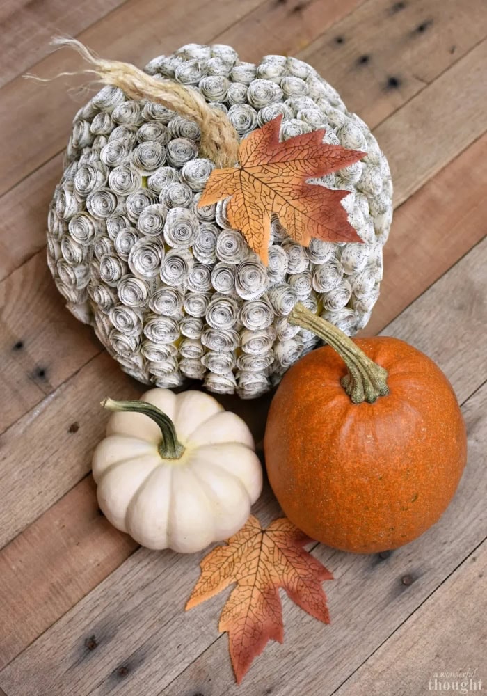 A very special looking pumpkin cover with tiny bookpage rose shapes, then decorated with an orange leaf.  It is sitting beside two smaller naatural pumpkins.  