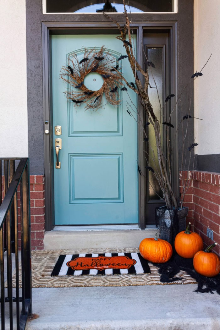 A Halloween front porch with a blue door.  The porch is decorated with a simple Halloween wreath, and branches decorated with bats.  There are pumpkins and a fun halloween door mat.