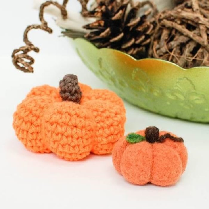A small crocheted pumpkin and a tiny felted pumpkin sitting beside a green bowl with dark brown foilage.